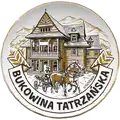 Ceramiczny magnes w kształcie talerza o średnicy 65mm w złotej ramce z brązowym nadrukiem, przedstawiający drewnяну архітектуру та кінний віз у Bukowina Tatrzańska, produkt turystyczny na sprzedaż hurtową B2B, POL-MP-229.