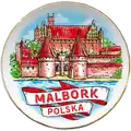 Ceramiczny magnes dekoracyjny w kształcie talerzyka o średnicy 65mm, ze złotym obramowaniem i kolorowym nadrukiem przedstawiającym Zamek w Malborku. Produkt turystyczny na sprzedaż hurtową B2B, POL-MP-221-2.