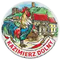 Ceramiczny magnes w kształcie talerza o średnicy 6,5 cm z kolorowym motywem koguta i panoramy Kazimierza Dolnego. Stylowy pamiątkowy upominek turystyczny dla hurtowej dystrybucji B2B, POL-MP-226.